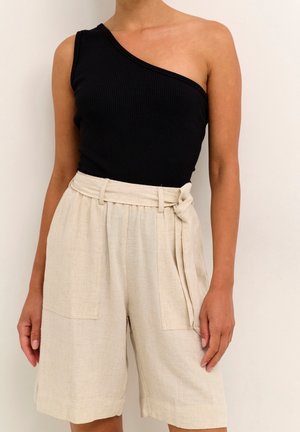 Kobieta ubrana w czarny top na jedno ramię i beżowe lniane szorty z wysokim stanem, z materiałowym paskiem wiązanym z boku.