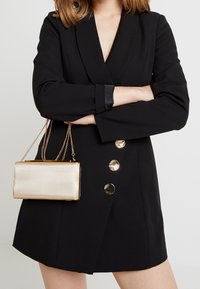 Il vestito blazer nero presenta un profondo scollo a V, maniche lunghe e bottoni metallici. Una clutch dorata e texturizzata con tracolla è tenuta in una mano.