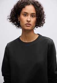 Sudadera negra con cuello redondo y puños acanalados, que presenta una textura suave y un diseño minimalista, llevada por una persona con cabello rizado.