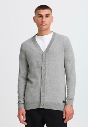 Grå cardigan med V-hals, frontknapper, ribbede mansjetter og kant. Brukt åpen over en hvit T-skjorte. Glatt tekstur. Uformelt design.