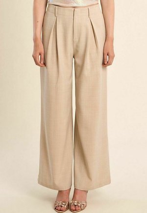 Pantaloni beige a vita alta e gamba larga indossati con sandali metallici intrecciati a punta aperta su uno sfondo chiaro.