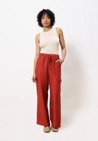 Lichtbeige geribde tanktop gecombineerd met losse, rode krinkel cargo broeken met zakken. Model staat tegen een witte achtergrond en draagt beige schoenen.