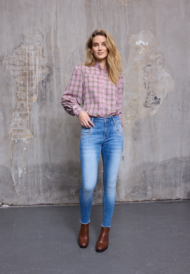 Camicia a quadri in tonalità rosa e verde, jeans skinny azzurri, stivaletti marroni, con sfondo di una parete grigia e testurizzata.