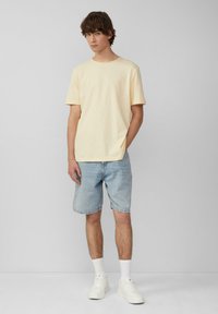 Lichtgele T-shirt met korte mouwen gecombineerd met denim shorts. Het model draagt witte sneakers en enkelsokken in het wit. Eenvoudige, casual uitstraling.