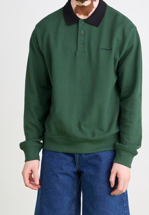 Grøn langærmet polo med en sort krave og tre knapper. Stoffet har et tekstureret mønster og er kombineret med blå vidde bukser i denim.