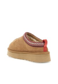 Brune ruskinds slip-on sko med shearling-fôr, rød og hvid broderet kant, tyk sål og rød løftestrop på hælen.