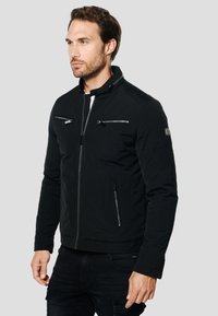 Mustast onast jakist, millel on kõrge krae, tõmblukuga taskud ja sile tekstuur. Omaduseks on keha kontuurimist järgiv lõige ja peened õmbluse detailid.