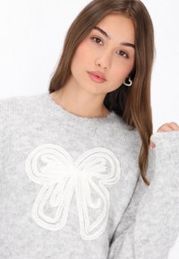 Pulover gri tricotat, cu un design proeminent de fundă albă în centru. Decolteu rotund și mâneci lungi cu croială ușor supradimensionată.