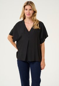 Svart V-ringad blus med korta, lösa ärmar, mjukt material och en subtil plissédetalj fram, kombinerad med mörka denimjeans.