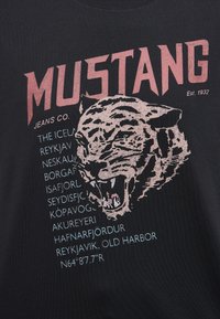 Černé tričko s grafikou tygří hlavy se zelenýma očima. Text zahrnuje "MUSTANG", "JEANS CO." a názvy islandských míst.