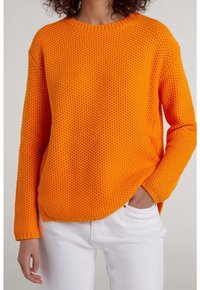 Maglione a maglia arancione brillante con motivo testurizzato, collo e polsini a coste, e vestibilità comoda. Indossato con jeans bianchi, mani nelle tasche.