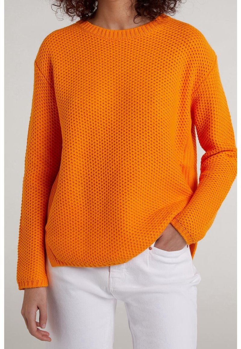 Maglione a maglia arancione brillante con motivo testurizzato, collo e polsini a coste, e vestibilità comoda. Indossato con jeans bianchi, mani nelle tasche.