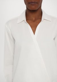 Valkoinen pitkähiha shirt klassisella kauluksella ja vinolla etuosa-alueella. Valmistettu sileästä kankaasta, jossa on minimaalinen yksityiskohta.