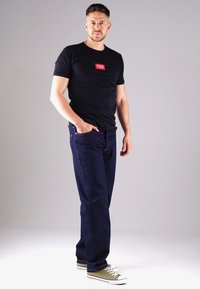 Schwarzes T-Shirt mit rotem Logo, kombiniert mit dunkelblauen Jeans und olivfarbenen Sneakers. Das T-Shirt hat kurze Ärmel; die Jeans sind im geraden Schnitt.