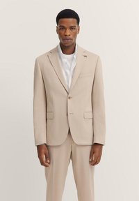 Beige maßgeschneiderte Anzugjacke mit Revers, Ein-Knopf-Verschluss, Fronttaschen, strukturierter Stoff, getragen über einem weißen Hemd.