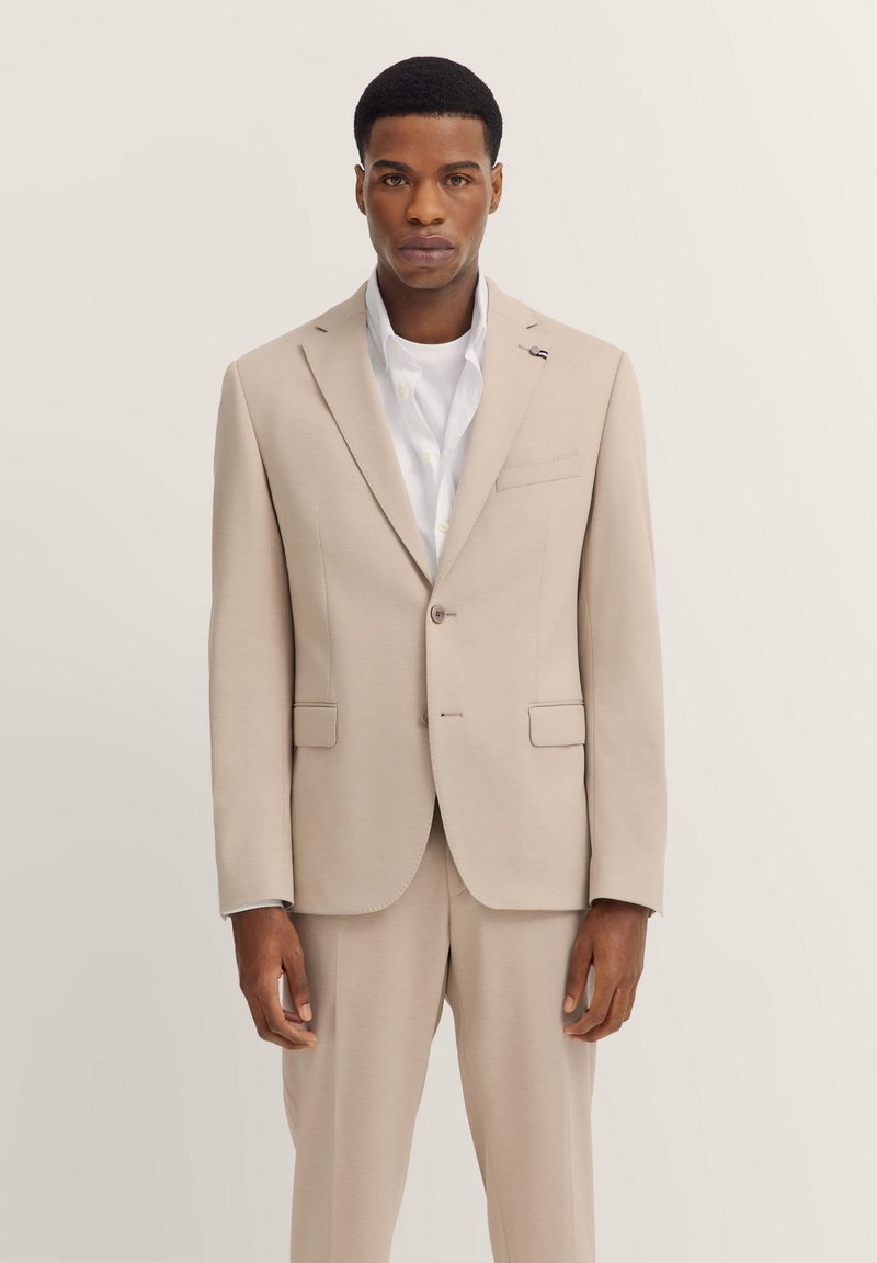 Beige maßgeschneiderte Anzugjacke mit Revers, Ein-Knopf-Verschluss, Fronttaschen, strukturierter Stoff, getragen über einem weißen Hemd.