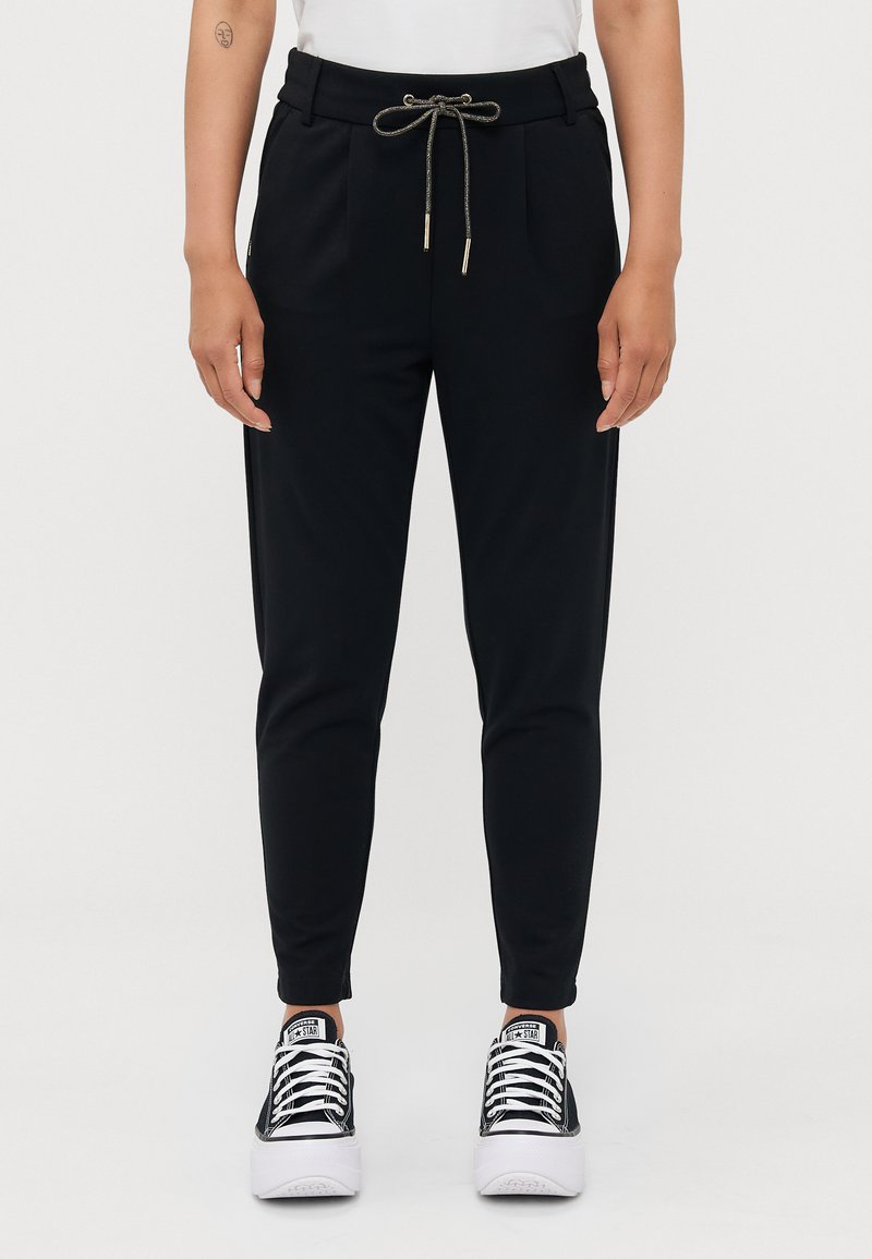 Pantalons de jogging noirs avec une taille élastique à cordon, dotés de poches latérales et de jambes fuselées. Le matériau a une texture lisse. Portés avec des baskets noires et blanches.