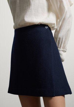 Woman wearing a dark navy high-waisted A-line skirt paired with a white long-sleeve blouse with buttoned cuffs.