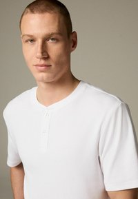 Camiseta henley de manga corta acanalada blanca con cuello redondo y tapeta de tres botones, mostrada sobre un fondo neutro.