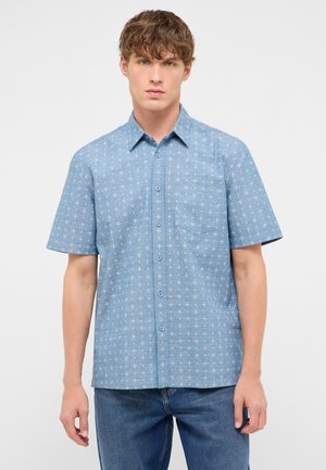 Junger Mann, der ein blaues Kurzarmhemd mit kleinem weißem Muster und blaue Jeans trägt, steht vor einem einfarbigen Hintergrund.