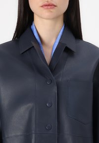 Navy leather jacket with a collared design, featuring a button front, single pocket, and a light blue striped collar visible underneath.
