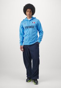 Helles blaues Kapuzenjacke mit Reißverschluss, ausgestattet mit "FRANCE" in Schwarz und einer französischen Flagge. Kombiniert mit lockeren, dunklen Jeanshosen und schwarzen Sneakers mit grünen Akzenten.