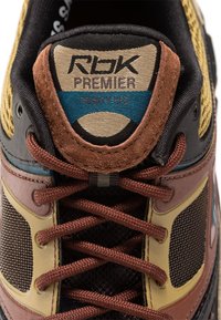 Tuviešais skats uz daudzkrāsainu sporta apavu mēli un šņorēm, kur uz auduma etiķetes redzama "RBK Premier Trinity RFS" zīmola nosaukums.