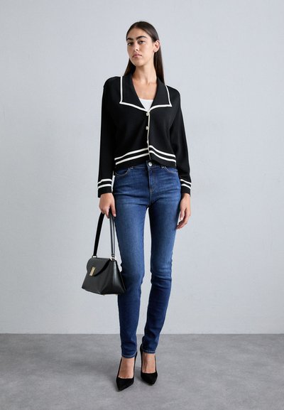 Svart kort cardigan med vit kant, tillsammans med blå tighta jeans. Modellen håller en svart handväska och bär spetsiga svarta klackar.