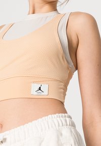 Zbliżenie na osobę noszącą beżowy krótki top z siateczki nałożony na jasnoszary podkoszulek bez rękawów oraz białe spodnie na sznurki.