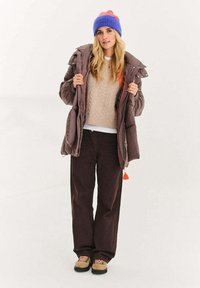 Puchowa, brązowa kurtka z pomarańczowymi akcentami, beżowy dziergany sweter, ciemnobrązowe spodnie, do tego niebiesko-pomarańczowa dziergana czapka i brązowe buty.