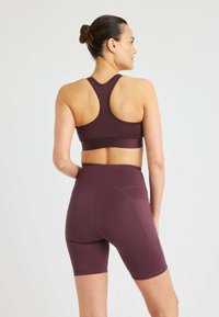 Donna con i capelli raccolti a bun, che indossa un reggiseno sportivo abbinato di un viola scuro e pantaloni ciclisti a vita alta, in piedi di spalle su uno sfondo chiaro e semplice.