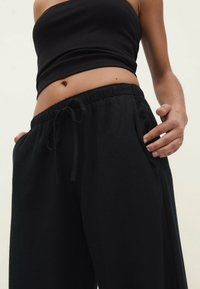 Pantalon de survêtement noir, ample, avec une taille élastique et un cordon de serrage. Texture douce et poches latérales pour plus de confort et de fonctionnalité.