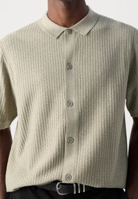 Korte mouwen, geborduurde blouse gemaakt van textuurstof met ribbels in lichtgroen, met een knoopsluiting aan de voorkant en contrasterende grijze knopen.