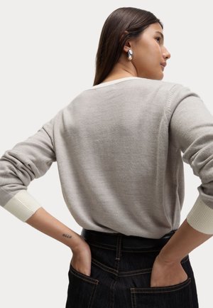 Lichtgrijze trui met geribbelde witte manchetten, ronde hals en zachte textuur, gedragen met donkere jeans, handen in de zakken, waarbij de achterkant zichtbaar is.