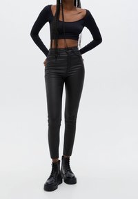 Schwarze Faux-Leder-Hose mit hoher Taille, schlanker Passform und glatter Textur, kombiniert mit einem langärmligen schwarzen Cropped-Top und schwarzen Kampfstiefeln.