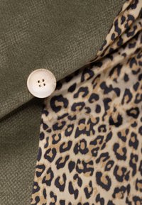 Olijfgroen stof met een gestructureerde afwerking, voorzien van een grote crèmekleurige knoop en een voering met luipaardprint in beige en zwarte tinten.