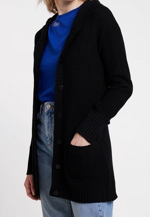 Personne portant un long cardigan noir en maille avec des boutons sur un T-shirt bleu et un jean bleu clair, main placée dans la poche du cardigan.
