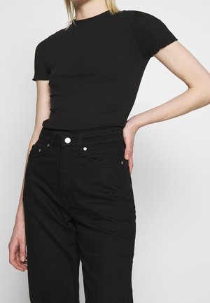 Personne portant un haut noir ajusté à manches courtes et un jean noir taille haute, avec une main sur la hanche, sur un fond uni.