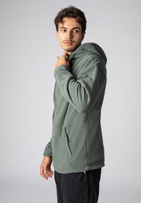 Grüner Fleece-Pullover mit Kapuze, ausgestattet mit einer seitlichen Reißverschlusstasche, verstellbarem Kordelzug und strukturierter Stoffqualität. Lange Ärmel, lockere Passform.