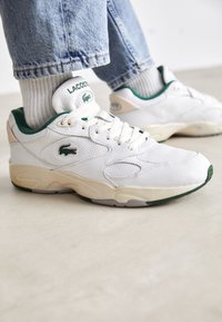 Białe skórzane sneakersy z zielonymi akcentami i perforowanymi detalami, z wyściełanym kołnierzem i teksturowaną podeszwą dla lepszej przyczepności.