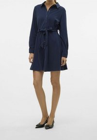 Vestito camicia blu navy realizzato in tessuto morbido, con chiusura a bottoni, maniche lunghe e vita legata. Abbinato a ballerine nere con punta.