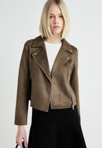Bruine faux suède motorjack met ritszakken, zilverkleurige accenten en een getailleerd ontwerp, gedragen over een lichte top en een zwarte rok.