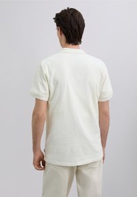 Tricou polo cu mânecă scurtă, culoare crem, material texturat, guler clasic, tiv drept. Purtat cu pantaloni deschiși la culoare, văzut din spate.