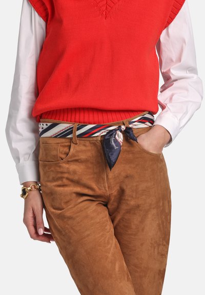 Sarkans adīts vestis virs baltas krekla, kopā ar brūnām zamšādas biksēm. Daudzkrāsains svītrainu šalli jostas detaļai. Zelta rokassprādze uz plaukstas locītavas.