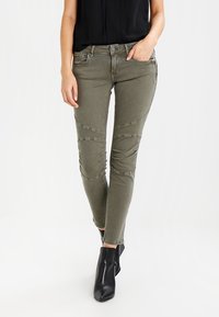 Olivově zelené džíny s úzkým střihem, s texturovanými detaily a švy. Doplněno černými kotníkovými botami.
