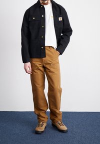 Giacca nera in tela con bottoni dorati, t-shirt bianca e pantaloni marroni. Indossa stivali beige con suole scure. Quattro tasche visibili sulla giacca.