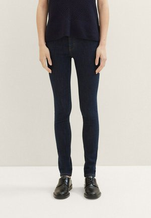 Hubené džíny v tmavém praní denimu, s hladkou strukturou a klasickým střihem, v kombinaci s černými botami pro elegantní vzhled.