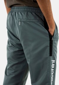 Heren trainingsbroeken in donkergroene stof, met een elastische tailleband, een ritssluiting achterzak en een wit logo langs de zijkant, met een gladde textuur.