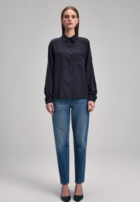 Granatowa koszula zapinana na guziki o teksturowanej tkaninie, z długimi rękawami i akcentami na ramionach; w połączeniu ze zwężającymi się niebieskimi dżinsami i czarnymi czółenkami z ostrym noskiem.
