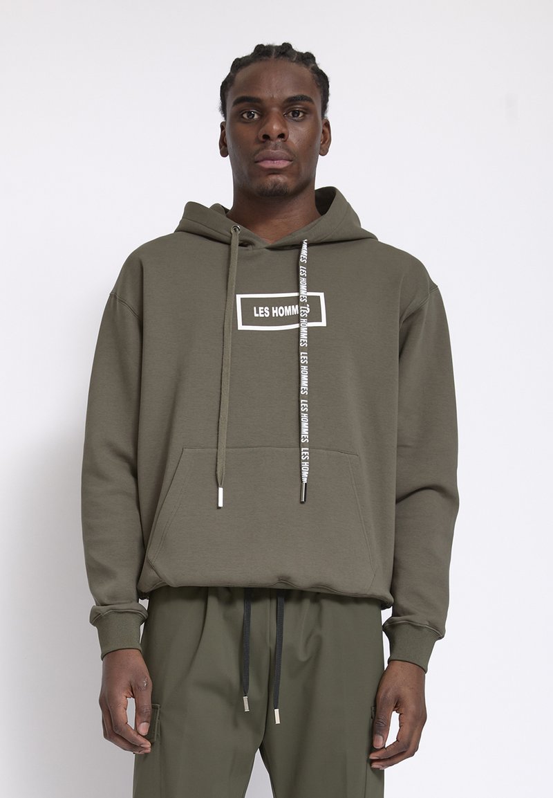 Jonge man met gevlochten haar, gekleed in een olijfgroene hoodie met de tekst "LES HOMMES" en bijpassende broek, staande tegen een effen achtergrond.